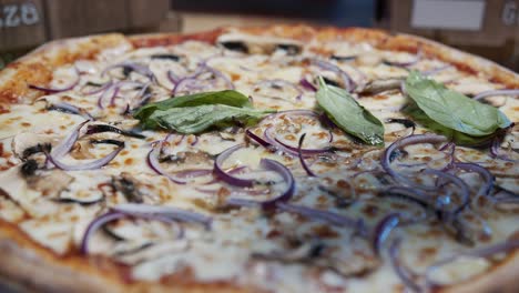 un primer plano de pizza fresca con champiñones, cebolla y queso mozzarella preparado sale del horno en una bandeja de metal hacia una superficie de madera rodeada de verduras frescas esparcidas sobre hojas de albahaca