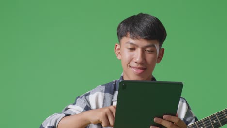 close up of asian teenager with guitar holding a tablet and pointing to side while standing on green screen background