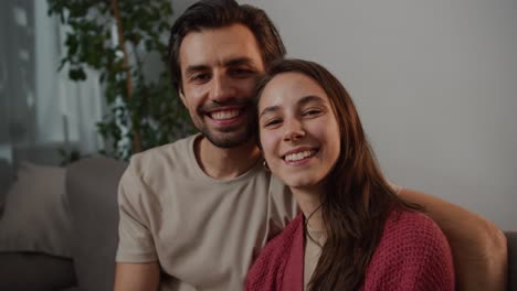 Close-up-portrait-of-a-happy-young-brunette-man-with-stubble-hugging-his-young-brunette-wife-in-a-red-sweater-and-posing-while-sitting-on-a-gray-sofa-in-a-modern-apartment