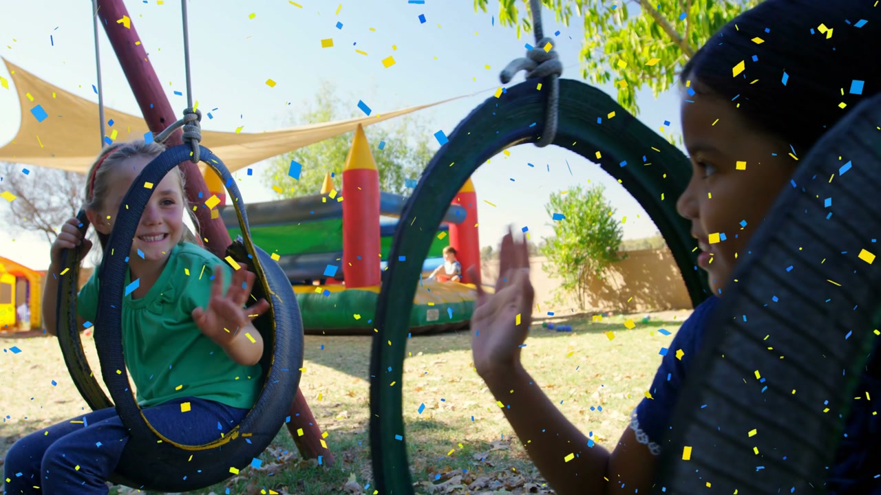 Two female children greeting on tire swings in yard as confetti appears camera panning bouncehouse