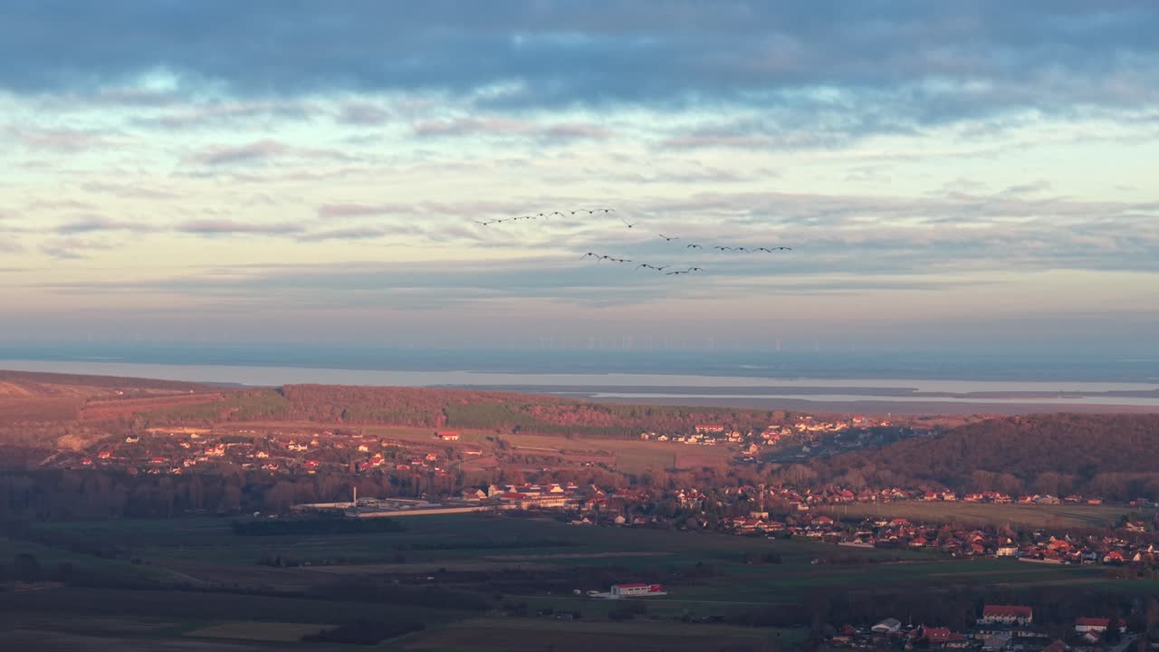Aerial view of a rural landscape illuminated by warm sunrise light, showing forests, and distant water. A flock of birds flies across the sky beneath layered clouds