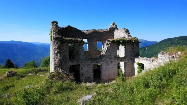 Casa de pedra em ruínas de dois andares com telhado desabado, aberturas de janelas retangulares vazias, alvenaria envelhecida, grama alta, pedras espalhadas e cristas de montanhas distantes sob um céu claro e luminoso.
