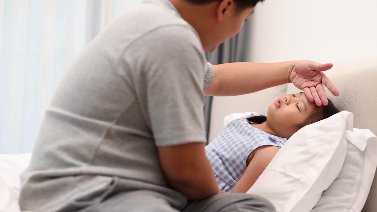 A father checks his daughter's temperature as she lies in bed, conveying care and concern in a softly lit room