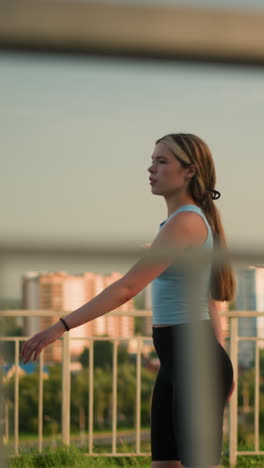 una joven patinando al aire libre en un día soleado, con una barandilla de hierro en primer plano, un paisaje urbano borroso de edificios altos, árboles verdes y el resplandor del atardecer en el fondo