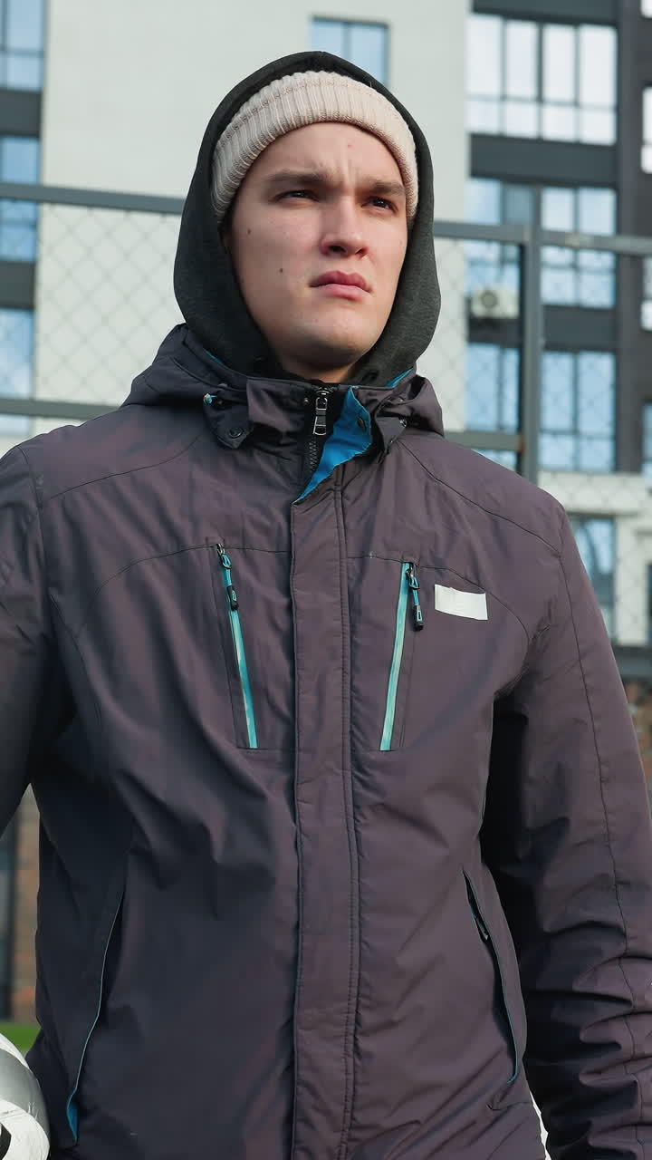 Young man in hoodie and beanie holding ball in right hand walks through bar fence with serious expression, tall office building in background