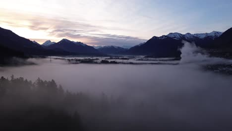 Drohnenumlaufbahn-Um-Nebligen-Kiefernwald-Mit-Sonnenaufgang-Und-Schneebedeckter-Naturberglandschaft-Im-Hintergrund