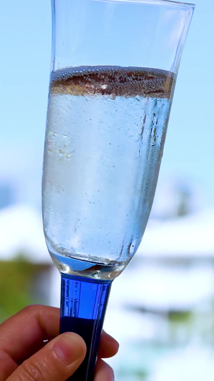 Una mano inclina lentamente un calice di champagne trasparente riempito di spumante frizzante contro uno sfondo esterno luminoso e soleggiato, evidenziando bollicine effervescenti e riflessi