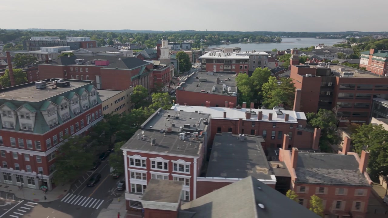 Ornate brick building with green mansard roof plus dormer windows anchors downtown corner, church clock tower rising above rooftops near harbor in Salem, Massachusetts. Aerial drone wide shot