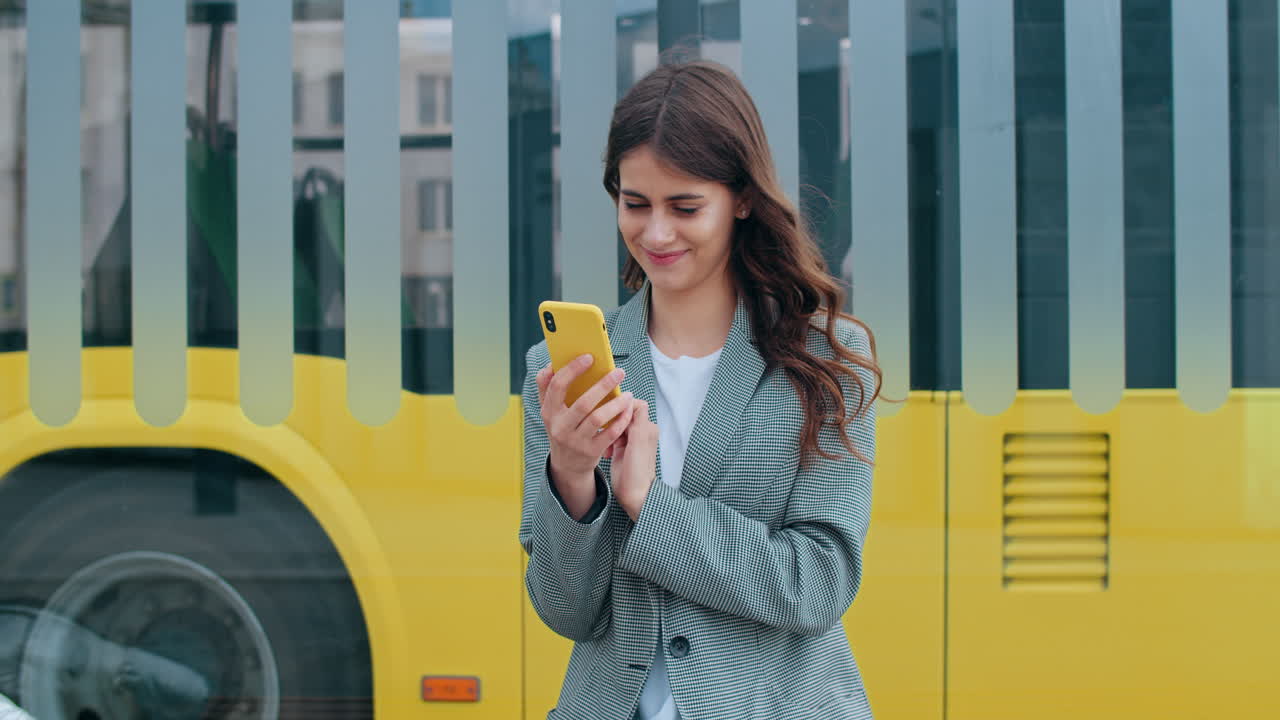 Young Woman Smiling While Using Her Smartphone Near a Bus