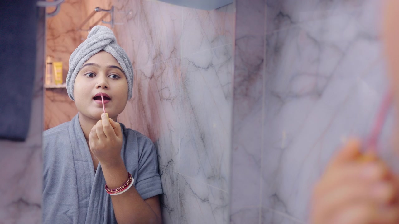 Indian woman applying lipstick in front of a bathroom mirror during her daily makeup routine.
