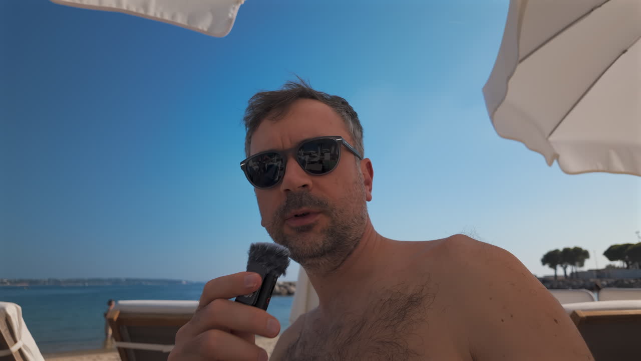 Man wearing sunglasses speaks into a small microphone under large white beach umbrellas with palm trees in the background