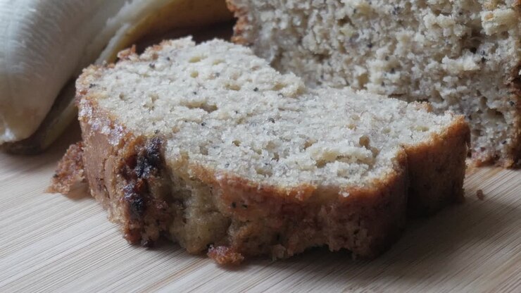 Left pan view of banana bread cut in half on a wooden board with a bananas in the back.