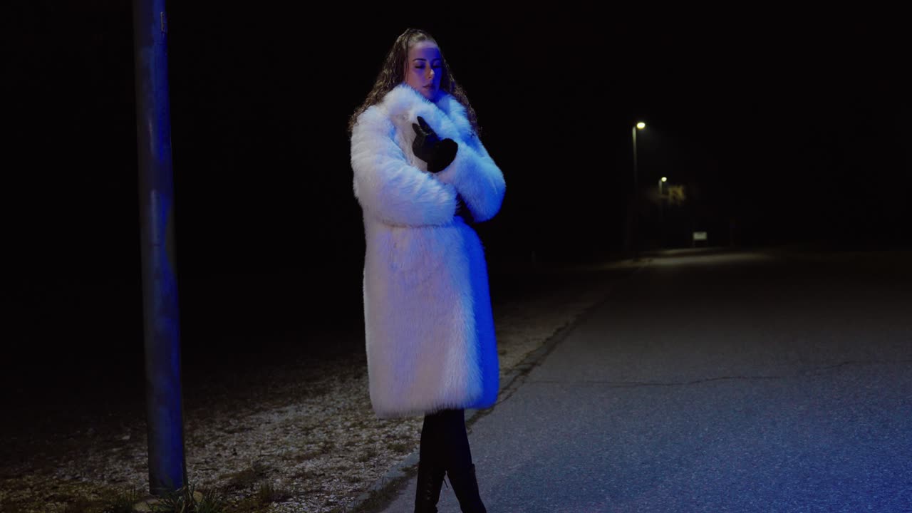 Foto notturna grandangolare di una donna in piedi da sola su una strada deserta, avvolta in un cappotto bianco e che si stringe per scaldarsi. Concept per freddo invernale, solitudine, vulnerabilità e atmosfera urbana tranquilla.