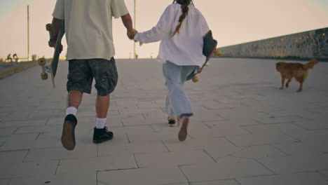 carefree teens strolling street back view. happy skaters holding hands walking