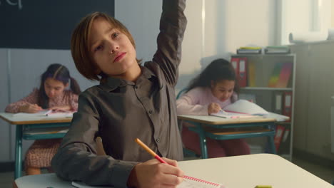 Boy-raising-hand-during-lesson-at-elementary-school.-Student-writing-in-notebook