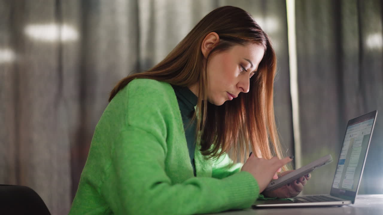 una mujer con un suéter verde desplazándose a través de su teléfono mientras trabaja en una computadora portátil en una mesa. capturado con una cámara de mano, destacando la multitarea y el trabajo digital moderno. trabajo remoto, tecnología