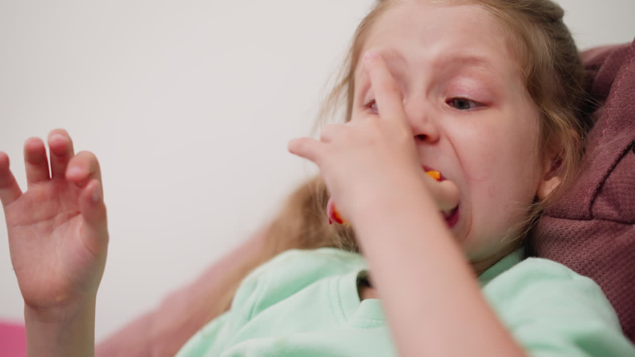 Cute girl in mint color cloth sitting on soft couch trying to take big bite from juicy apple holding it with both hands showing focus and determination indoors cozy playful environment