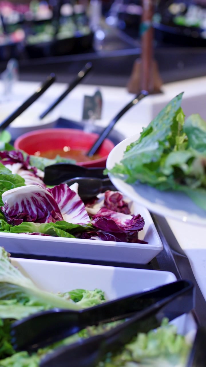 Primo piano di una donna che usa le pinze a un buffet di insalate, selezionando verdure a foglia sotto una luminosa illuminazione interna.