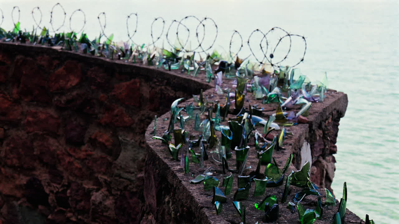 Close up of multiple broken glass shards and barbed wire loops along the edge of a brick wall with a blurred view of the sea