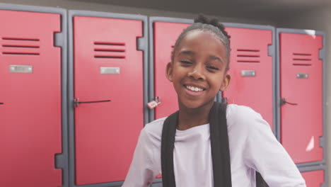 smiling african american girl with backpack standing in front of red lockers at school