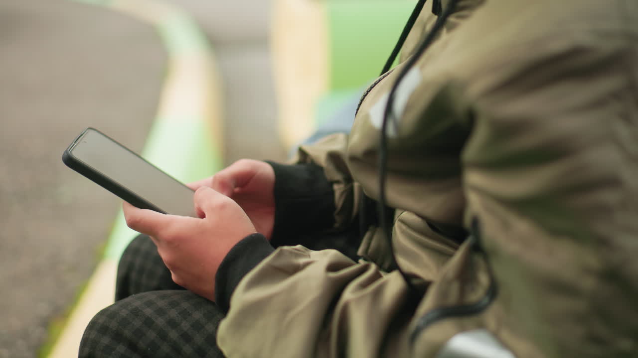Close up rear view of little kid in jacket holding smartphone with both hands, fully engaged in phone operation while sitting outdoors, showing focus and concentration on digital device