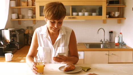mujer mayor usando el teléfono móvil mientras desayuna en la cocina