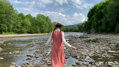 retro styled young woman in straw boater hat and long dress walking on river rocks near forest. romantic girl. natural scenic landscape. summer vacation. vintage aesthetic lifestyle. 4k video