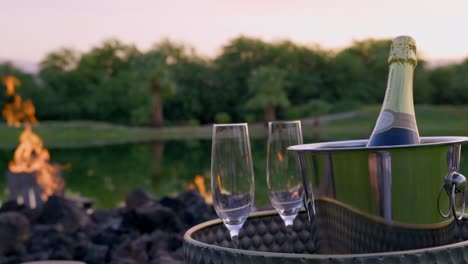 A bottle of Champagne inside a metal bucket with two glass cups near a bonfire and lake, Close up static