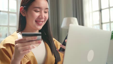 asian woman sitting on couch in living room holding credit card while using smartphone and laptop shopping online