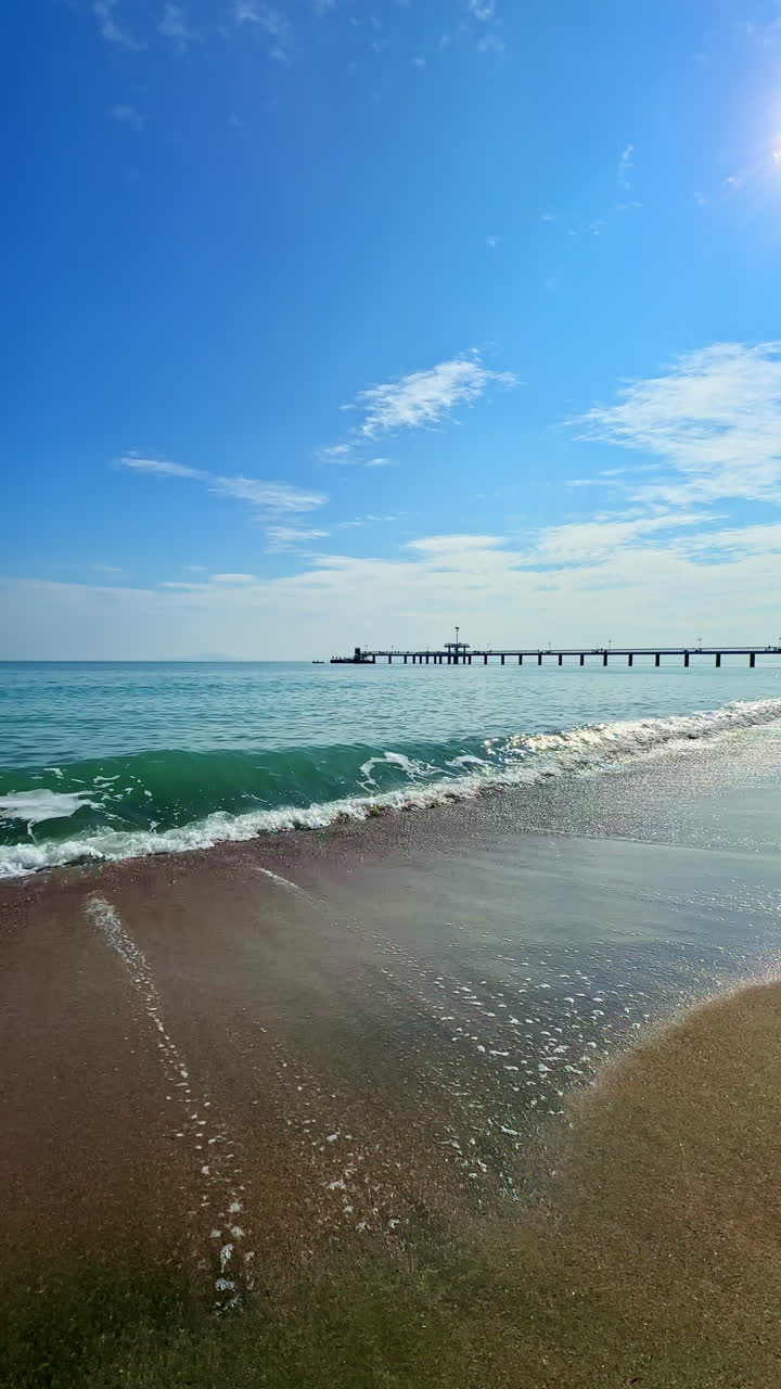 Waves gently lap at the serene beach as a distant pier stretches into the clear blue sky on a warm sunny day