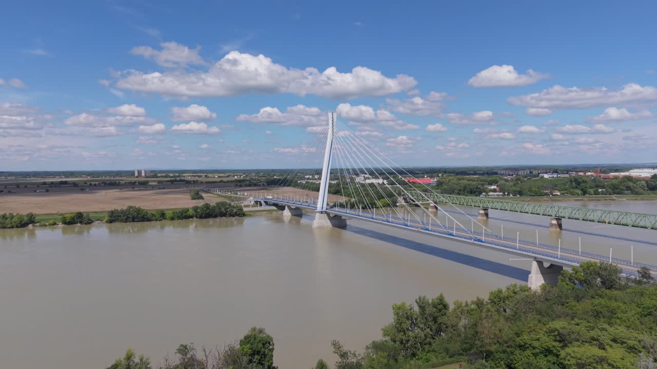 A modern cable-stayed bridge stretches across a wide river on a sunny day with clouds. The structure is set against a rural and urban landscape, highlighting the blend of infrastructure and nature