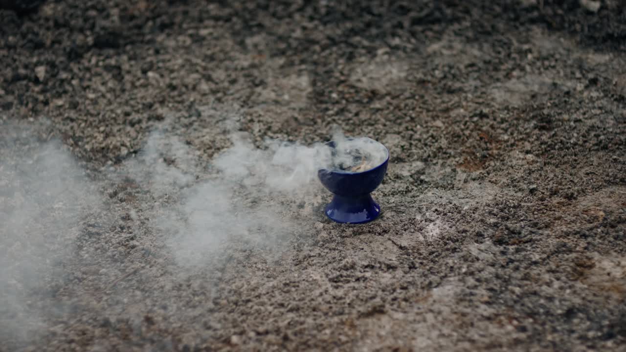 Sinisessä keraamisessa kulhossa palava salvia harmaalla, tuhkaisella alustalla, kun henkilön käsi tulee hetkeksi kuvaan oikeasta reunasta ja valkoinen savu leijuu sivuttain ilmassa