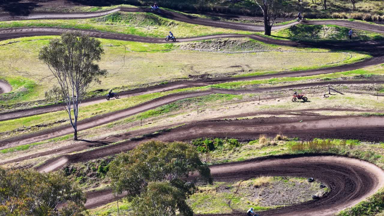 Multiple riders navigate a winding dirt motocross track surrounded by bushland, captured in bright daylight from a high, stable aerial drone perspective