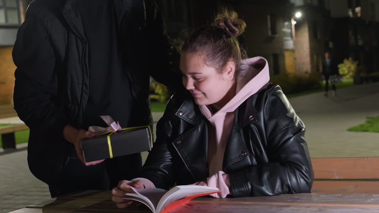 Lady in leather jacket and hoodie seated outdoors at wooden table reading book while husband surprises her by presenting wrapped gift under soft night lighting in cozy urban atmosphere