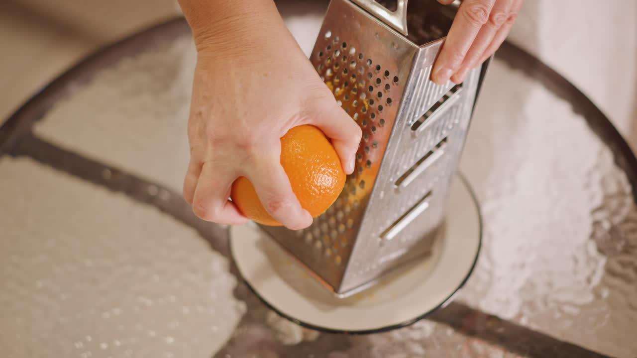 Primo piano di mani caucasiche che grattugiano una scorza d'arancia sopra un piatto con una grattugia di metallo, un movimento energico verso il basso che rilascia oli essenziali di agrumi e un aroma brillante, preparazione di guarnizioni per dessert con il calore accogliente della cucina.