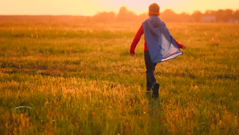 Junge-Im-Superheldenkostüm-Und-Mit-Maske-Rennt-Bei-Sonnenuntergang-Träumend-Und-Phantasievoll-über-Das-Feld.