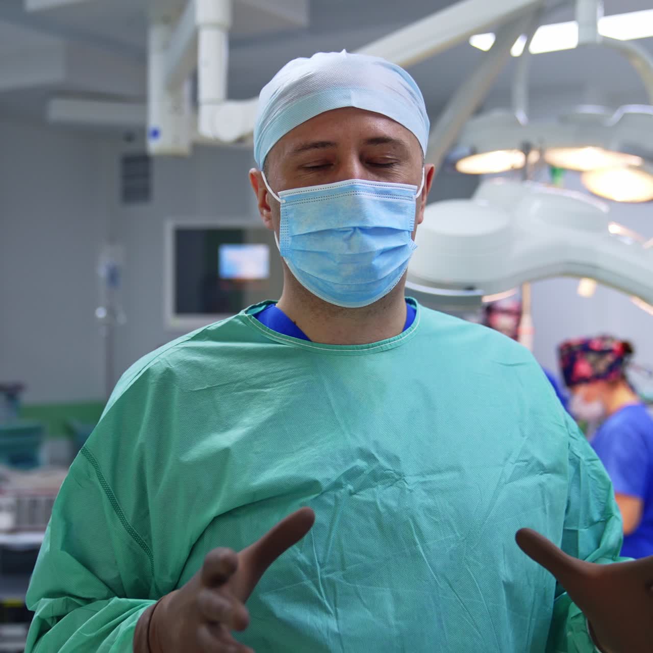 Professional surgeon in uniform, cap, mask and beige latex gloves talking to camera. Close up. Medical staff at the operational table at backdrop