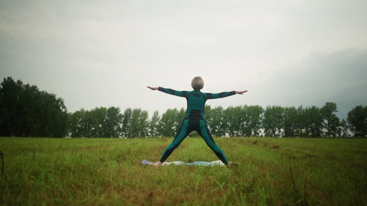 Back view of woman in green-black suit standing on yoga mat practicing triangle pose with arms wide open and legs apart, set against cloudy skies in a peaceful grassy field surrounded by nature