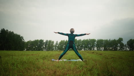 back view of woman in green-black suit standing on yoga mat practicing triangle pose with arms wide open and legs apart, set against cloudy skies in a peaceful grassy field surrounded by nature