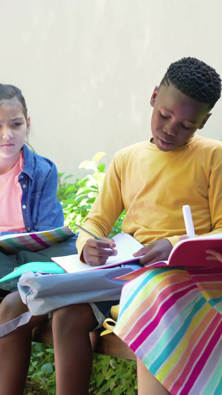 Vertical video: Adjusting boy in yellow tee head-down over notebook on backpack writing at school
