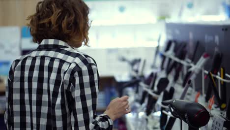 Young-cheerful-positive-woman-in-plaid-walks-choosing-curling-iron-in-household-appliances-store,-taking-one-from-the-shelf-and-examines-in-hands.-Slow-motion