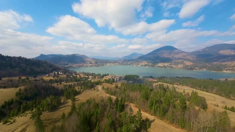 Impresionante-Vista-Aérea-Sobre-El-Lago-Colibita-En-Un-Hermoso-Día-Soleado,-Junto-Al-Lago-Rodeado-De-Bosques-Verdes-Y-Casas-Rurales-Dispersas-Con-Un-Hermoso-Fondo-De-Los-Cárpatos