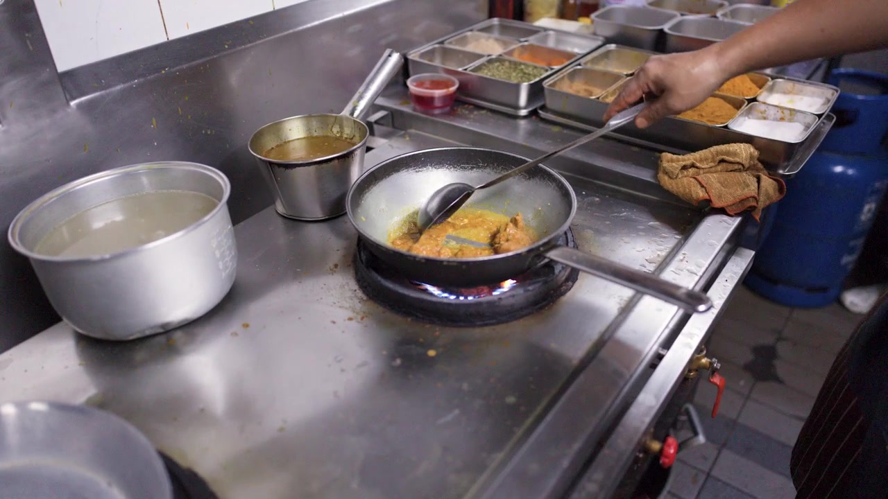 Chef asiático prepara comida em uma cozinha comercial usando uma panela de metal e fogão a gás.