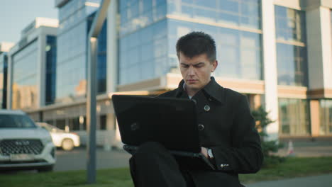 investor in black coat seated outdoors operating laptop with leg crossed, appearing deeply focused, urban background includes parked cars, modern office buildings, and open public space