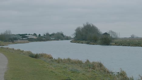 Toma-Panorámica-En-Un-Día-Gris-Nublado-En-Medio-Del-Invierno-Inglés-De-Un-Río-Con-Un-Camino-Y-Un-Corredor,-Ely-Inglaterra