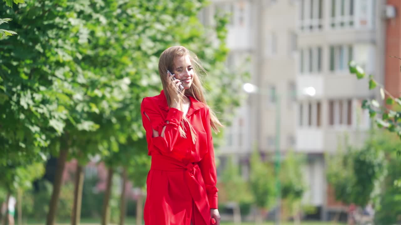 Gorgeous long-haired woman in red dress. Attractive lady talking the mobile phone on the city background in a bright summer day. Slow motion.