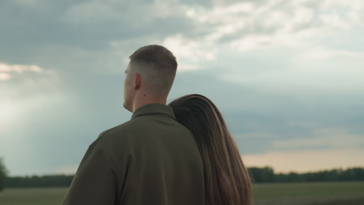 man and woman stand at edge of green field and cloudy sky as woman rests head on husband shoulder while he lifts arm pointing toward horizon conveying love comfort and hopeful future vision