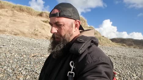 bearded man wearing baseball cap backwards looking down over sunny beach shoreline hunting and foraging