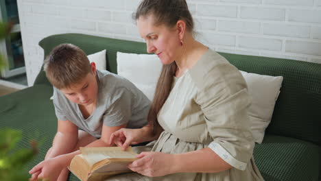 woman flipping book page while son in grey shirt sitting beside her attentively looking into old open book on green couch with white pillows, indoor cozy home setting, christmas tree slightly blurred