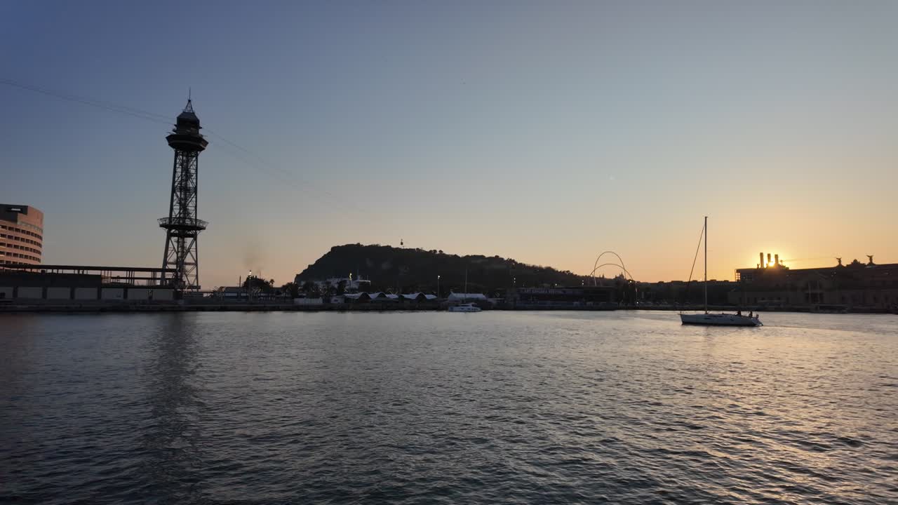 Barselona limanı üzerinde gün batımı manzarası, Port Vell teleferik kulesi, Montjuic tepesi ve sakin sularda seyreden bir yelkenli.